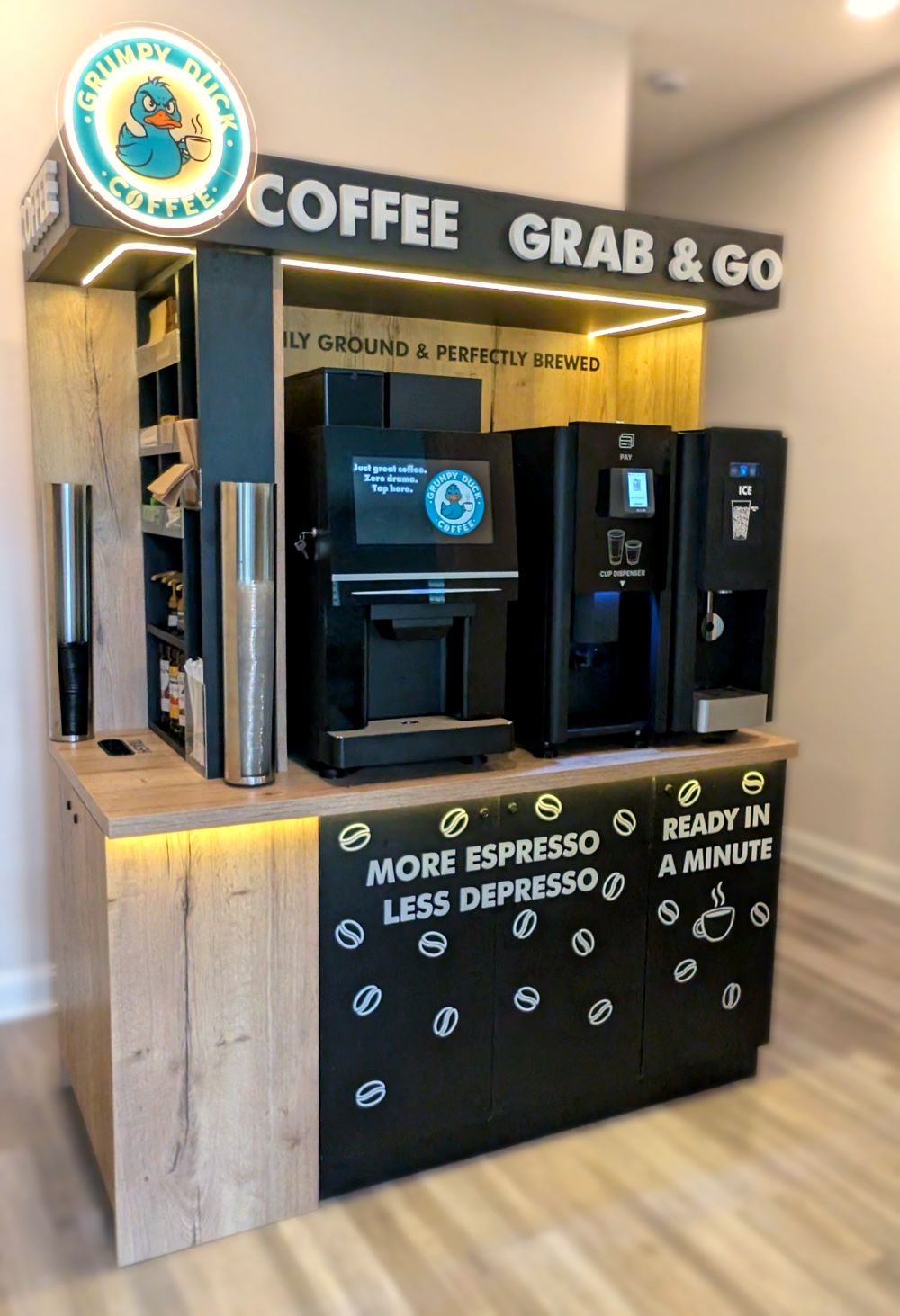 The Grumpy Duck coffee kiosk with a lit Grumpy Duck sign, coffee machine, cup dispenser and ice maker in a black and oak cabinet